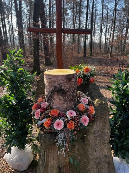 Holzkreuz im Wald mit Blumenkranz